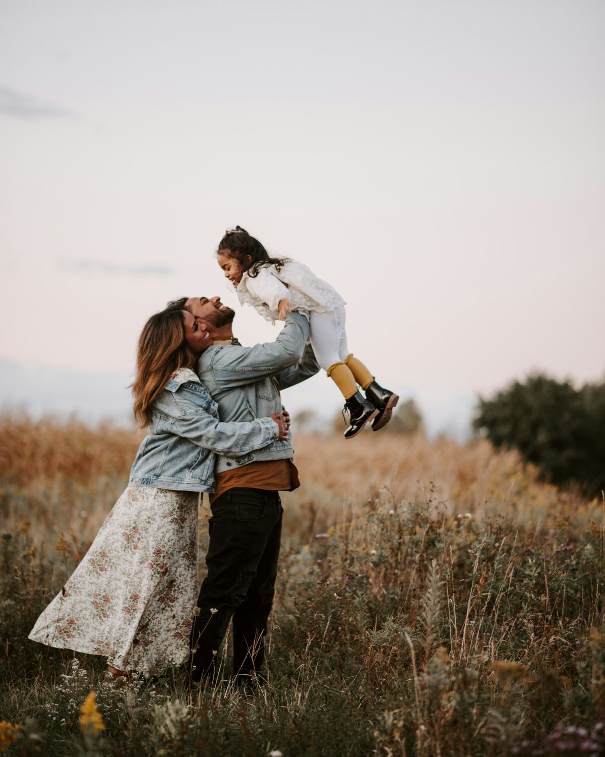 A father lifting his laughing daughter into the air in a golden field while her mother holds him.