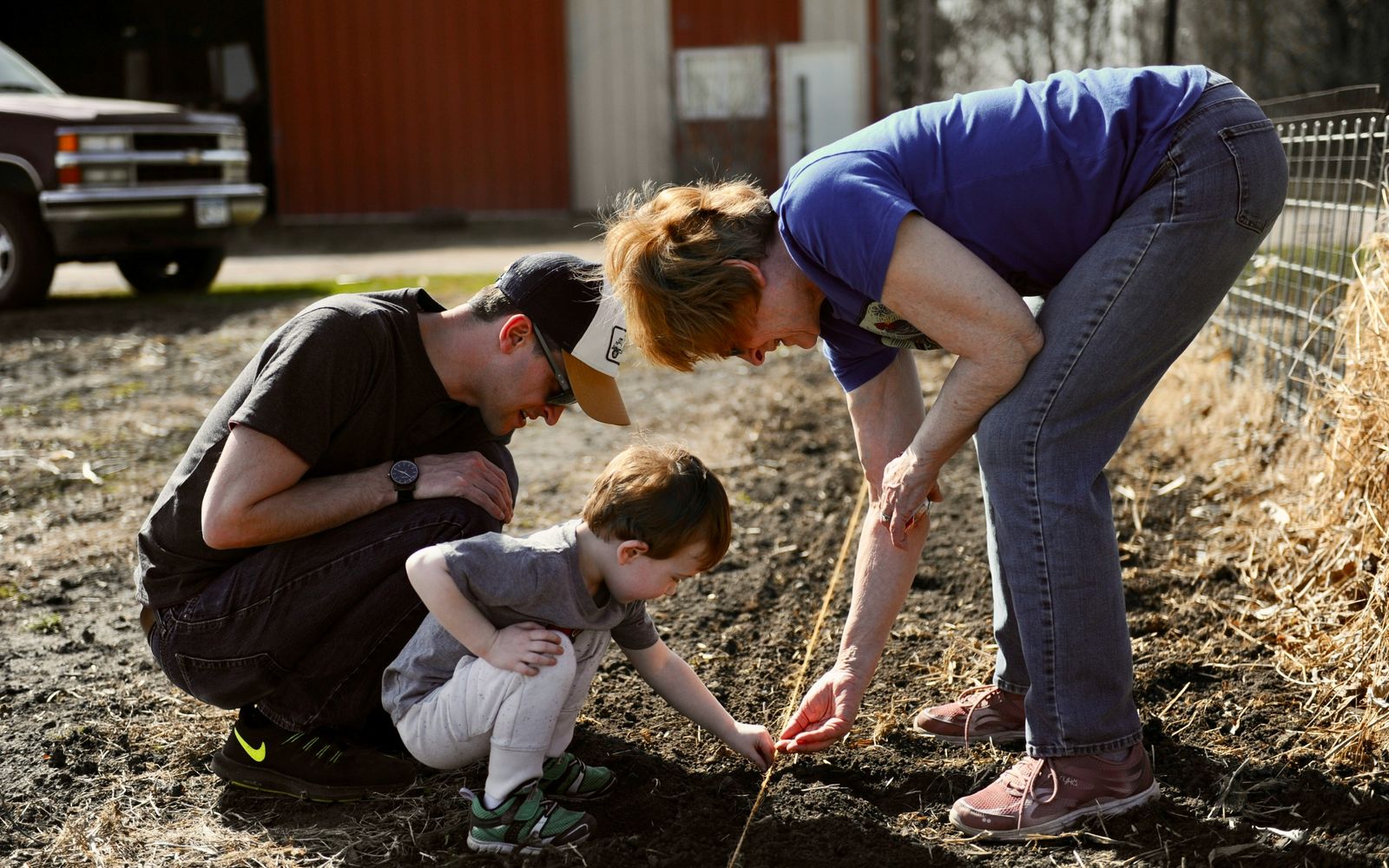 A grandmother and father showing a toddler how to plant seeds in a garden row.